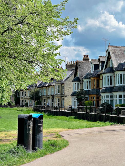 In the foreground, two black recycling bins are positioned on a paved pathway adjacent to a grassy area, with a large leafy green tree on the left side of the image. Behind the bins, there is a row of