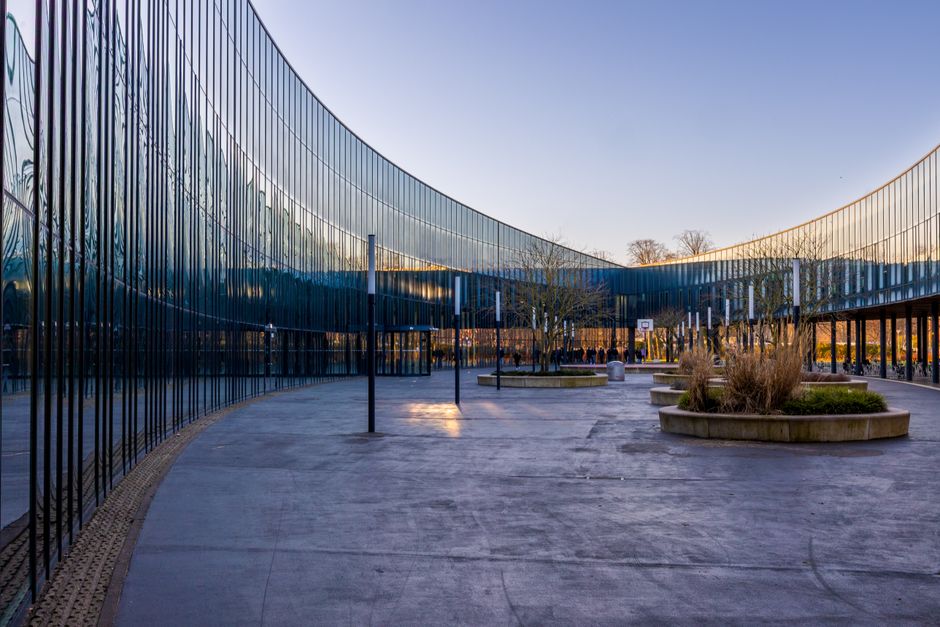 A tall modern office building with a glass facade and visible interior lights, situated behind a row of green trees and shrubs on a clear day. The building features multiple floors with exterior balco