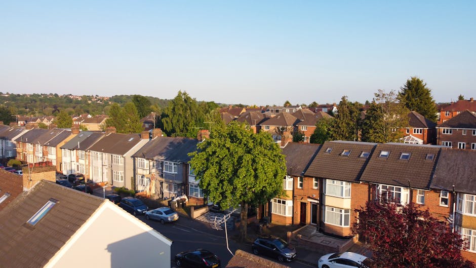 An aerial view of a residential neighborhood showing closely spaced terraced houses with small back gardens and driveways, along with some detached and semi-detached homes. The scene includes an adjac
