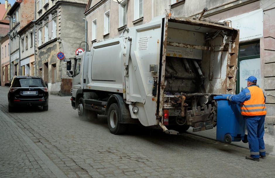 A waste collection worker wearing a high-vis vest and dark clothing is operating a bulk waste collection vehicle on a city street during early evening or late afternoon. The worker is facing away from