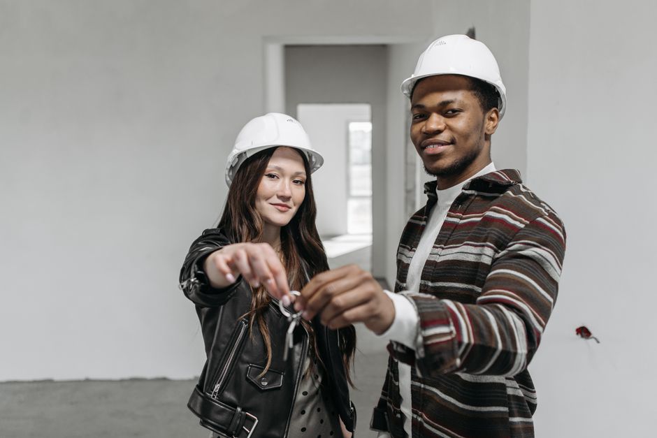 Inside a residential property, two movers wearing white safety helmets are engaged in a moving process. The female mover, with long dark hair and a black leather jacket, is handing over a set of keys