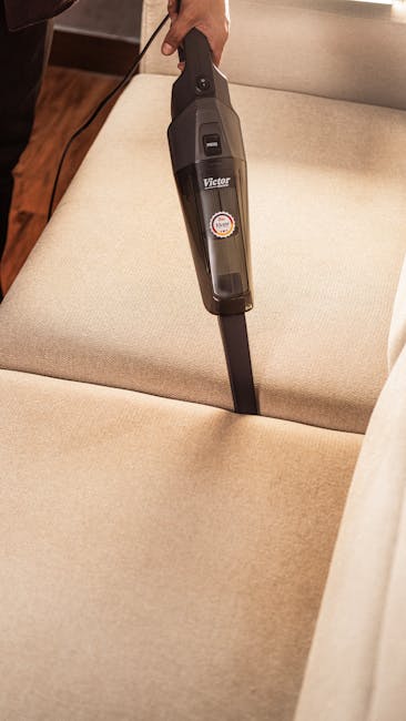 Close-up image showing a person using a handheld vacuum cleaner to clean a beige fabric sofa inside a home. The vacuum, labeled Victor, is held by a hand, with the nozzle pressed onto the sofa's cushi