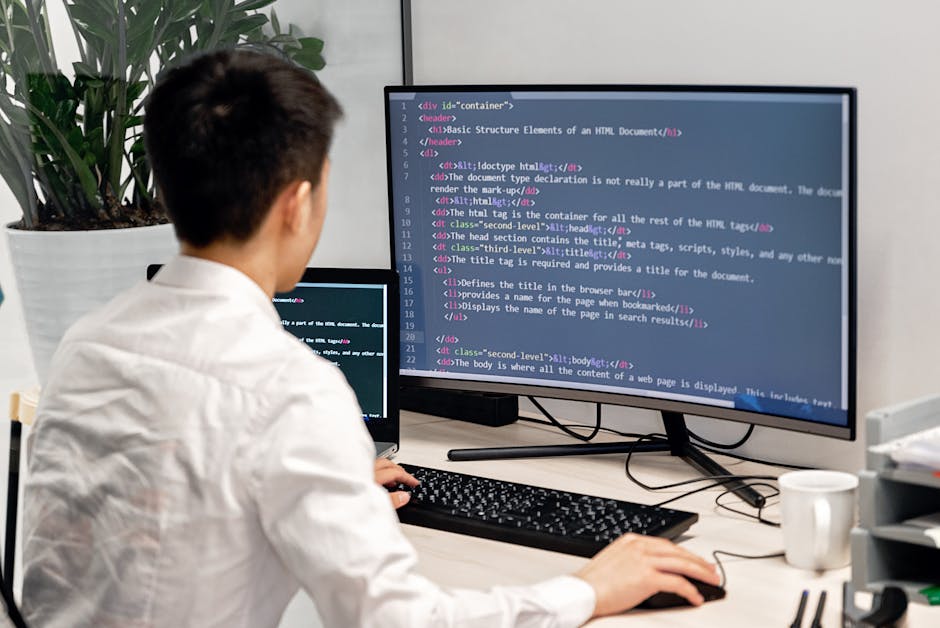 A person with short dark hair and wearing a white shirt sits at a desk in an office environment, working on a computer with dual monitors. The primary monitor displays a webpage with HTML code in a te
